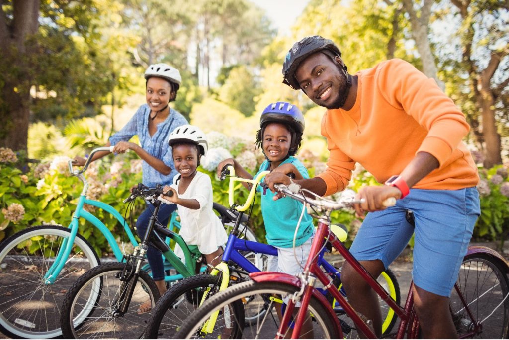 Mom, Dad, and two children smile at the camera while on bikes at the park on a sunny day