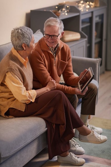 Older couple wearing matching brown slacks and sweaters laughs while sitting on a couch holding a tablet. 