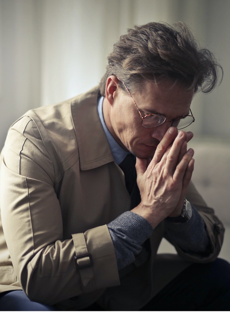 Man wearing glasses and a tan coat holding his hands to his face in exhausted contemplation