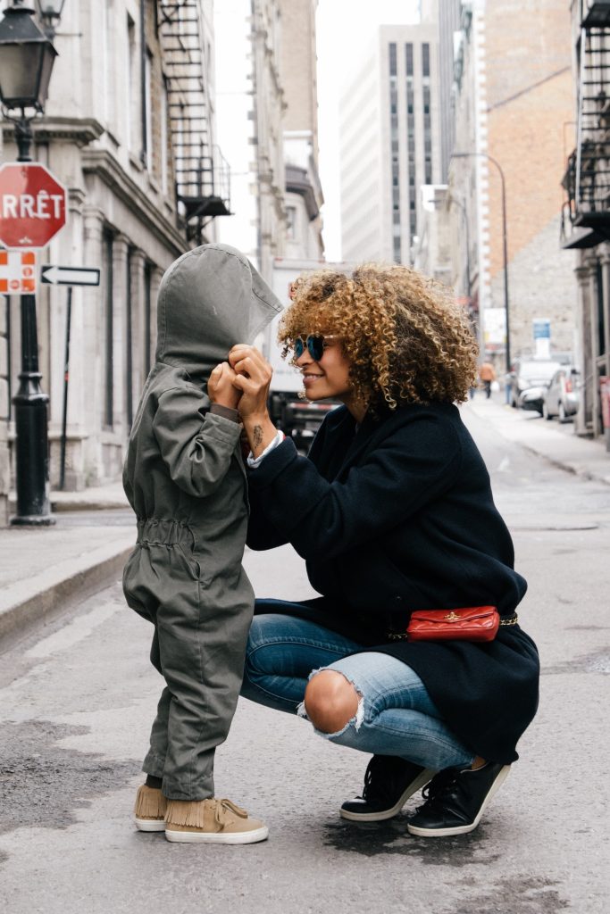 Mother with curly hair crouches down to height of her young child, holding hands on a city street.