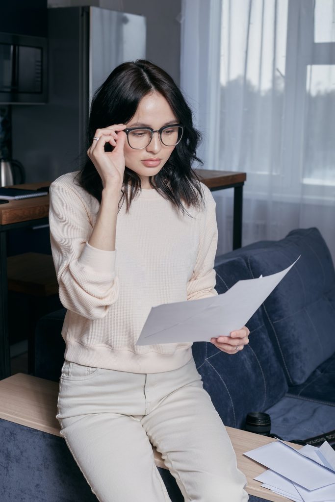 Woman with dark hair and glasses wearing a light tan sweater and pants reading papers held in her left hand