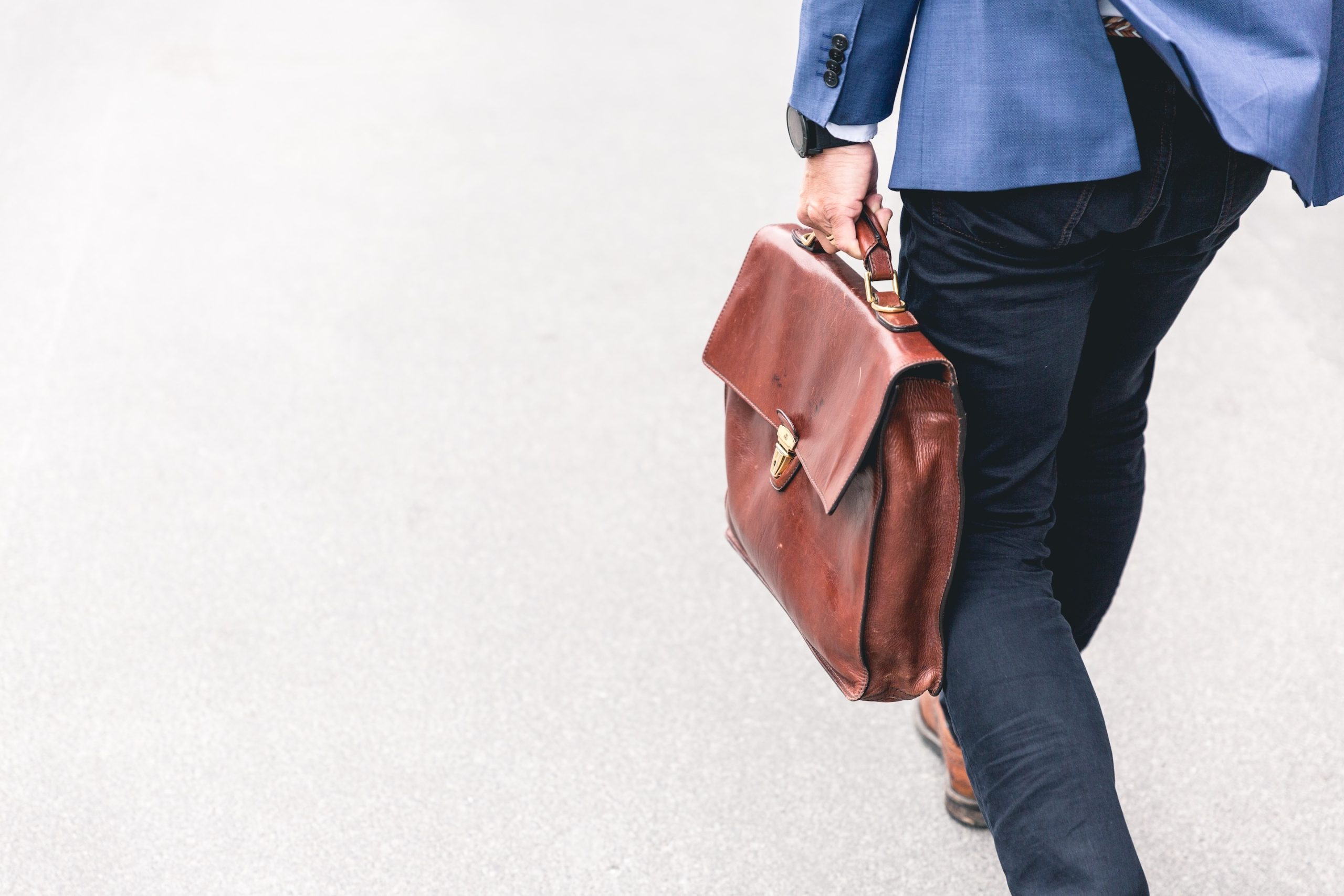 Man wearing a blue suit walking away with a brown suitcase in his hand.
