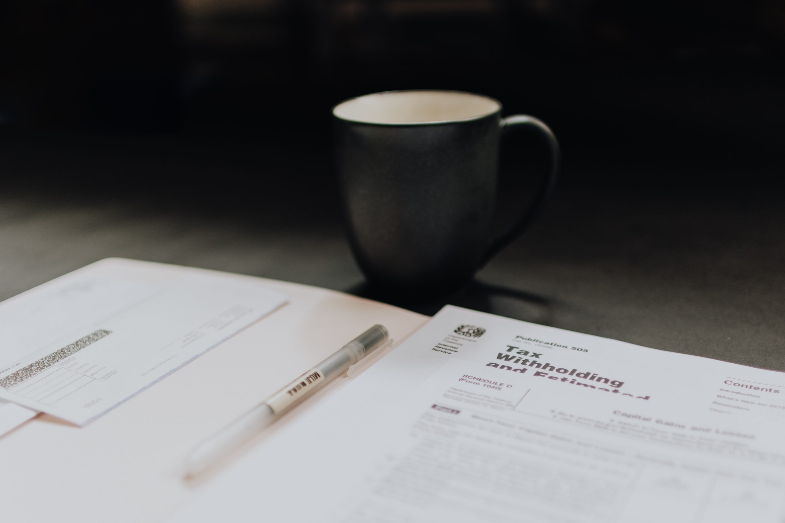 Tax forms spread out on a table with a pen on top. A black coffee cup sits in the background.